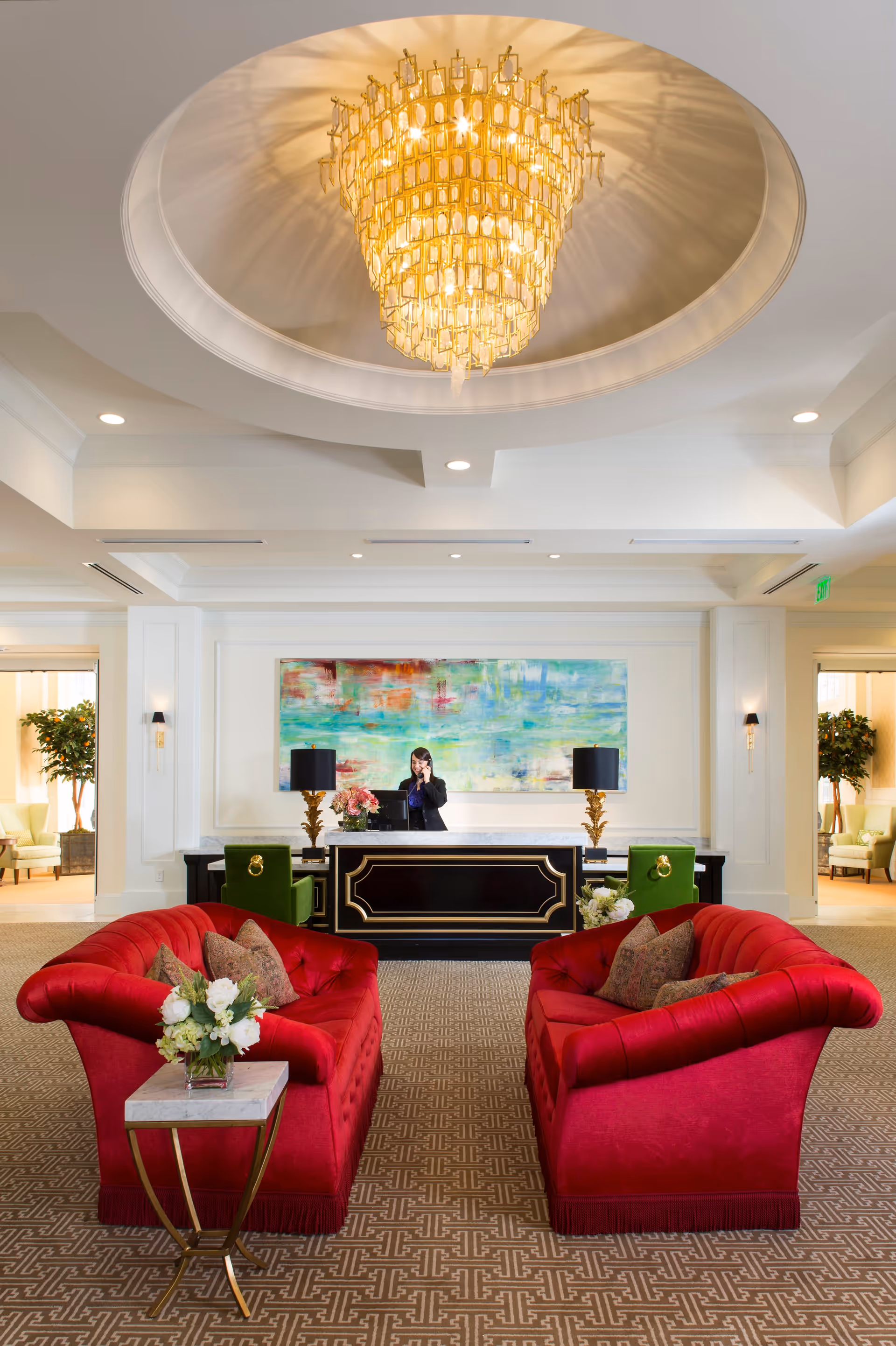 A luxurious reception area with two red velvet sofas facing each other, a small side table with a flower arrangement, and a large black and gold reception desk with a woman speaking on the phone behind it. The room features a large, ornate chandelier hanging from a recessed ceiling and a colorful abstract painting on the wall behind the desk. There are green chairs and plants visible in adjacent rooms.