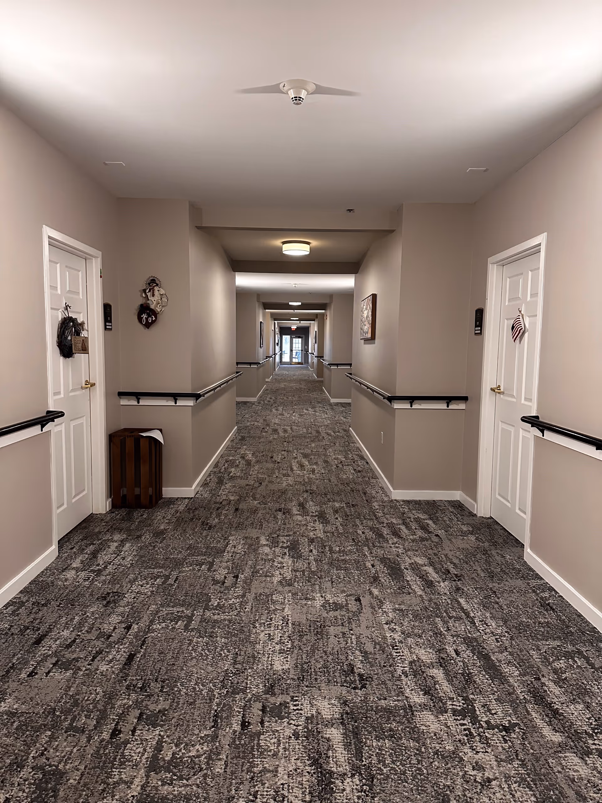 A long, carpeted hallway in a senior living facility with beige walls, white doors on both sides, handrails along the walls, and ceiling lights. Decorations hang on some doors and a window is visible at the far end of the corridor.