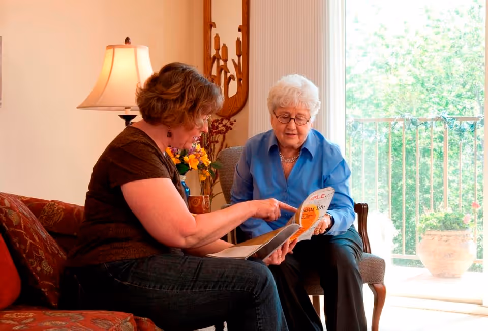 Two women sitting in a living room area, one younger and one elderly, looking at a booklet together. The room has a lamp, flowers, and a large window with a view of greenery outside.