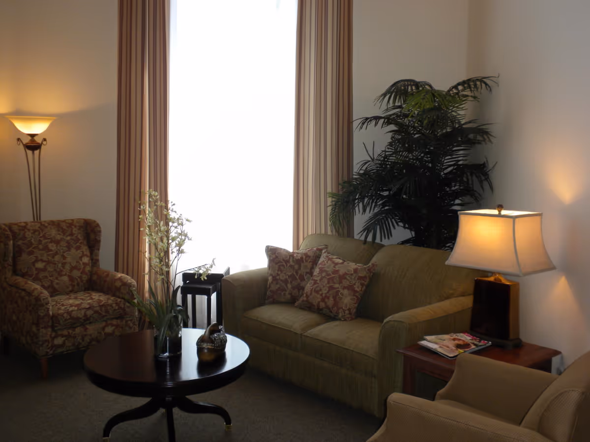 Cozy living room with a sofa, patterned armchairs, coffee table, lamps, and large potted plant by a curtained window.