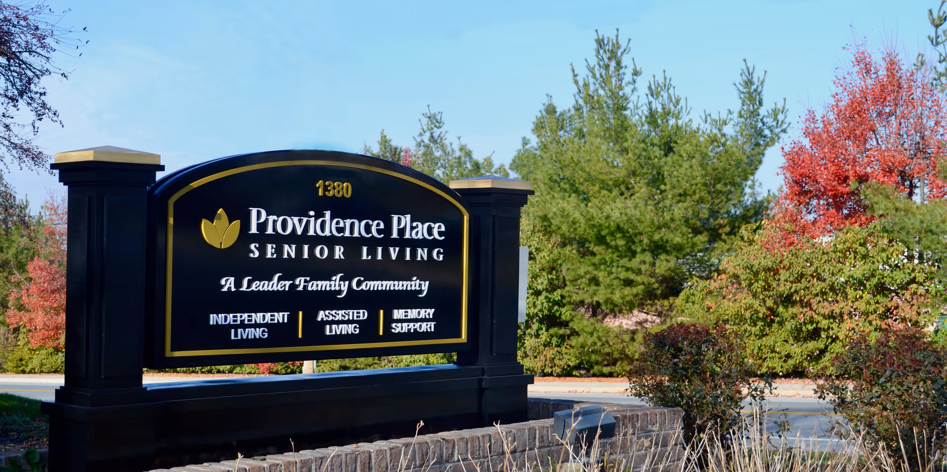 Outdoor view of a large black and gold sign for Providence Place Senior Living, located at 1380. The sign mentions it is a Leader Family Community offering Independent Living, Assisted Living, and Memory Support. The sign is surrounded by trees with green and red foliage under a clear blue sky.