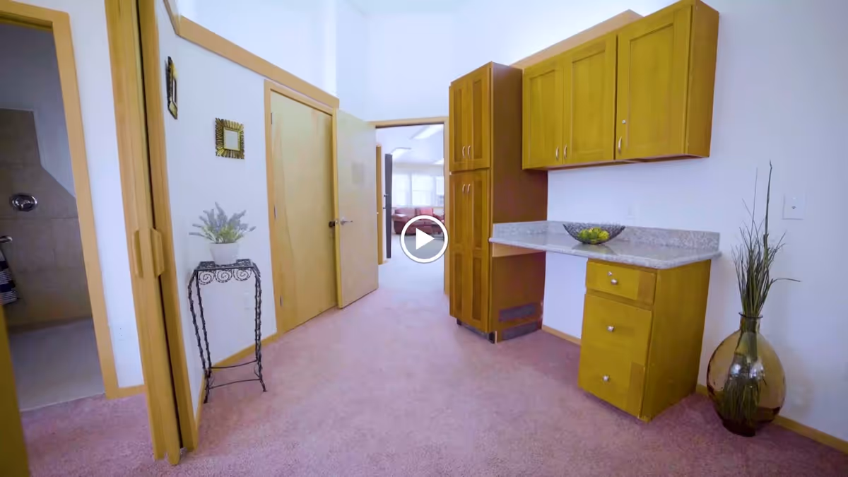 Interior view of a senior living facility hallway with light wood doors and cabinetry. There is a small countertop with drawers and cabinets, a decorative plant in a glass vase on the floor, and a small table with a potted plant. The hallway carpet is light pink, and a bathroom is visible through an open door on the left.