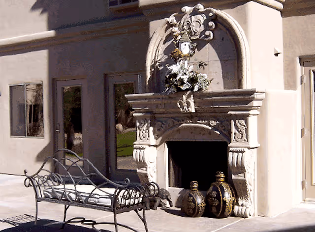 Outdoor patio area featuring a decorative stone fireplace adorned with a floral arrangement and ornamental vases at its base. In front of the fireplace is a metal bench with a white cushion. The background shows windows and doors of the building.