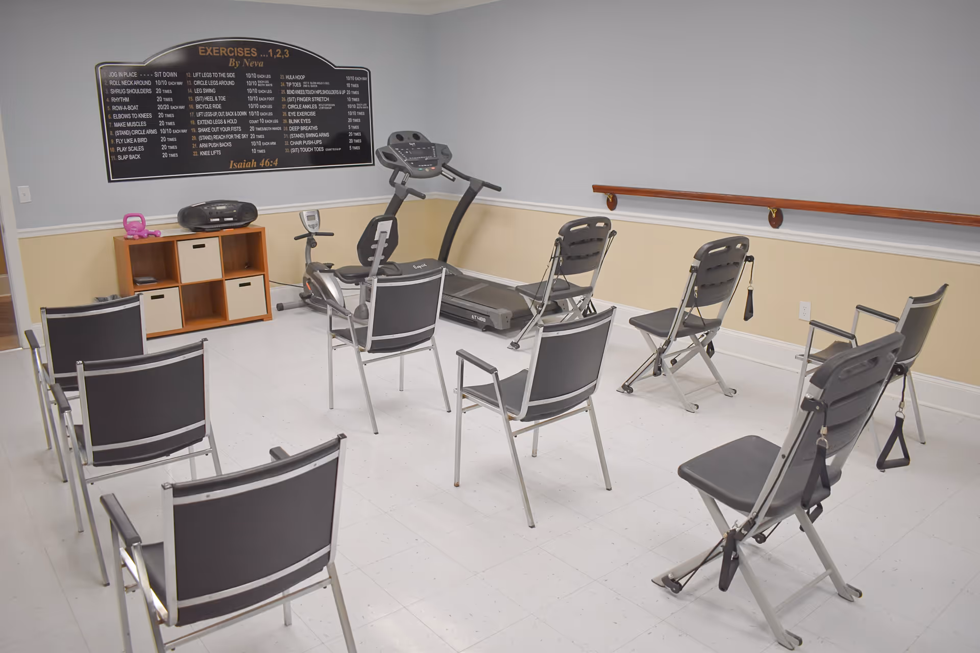 A small exercise room with eight black and silver chairs arranged in a semicircle facing a treadmill and a stationary bike. On the wall above a wooden shelf with a radio and a pink kettlebell is a large black exercise instruction board titled 'EXERCISES ...1, 2, 3' with various exercise steps listed. The room has light-colored walls with a wooden handrail on one side and a white tiled floor.