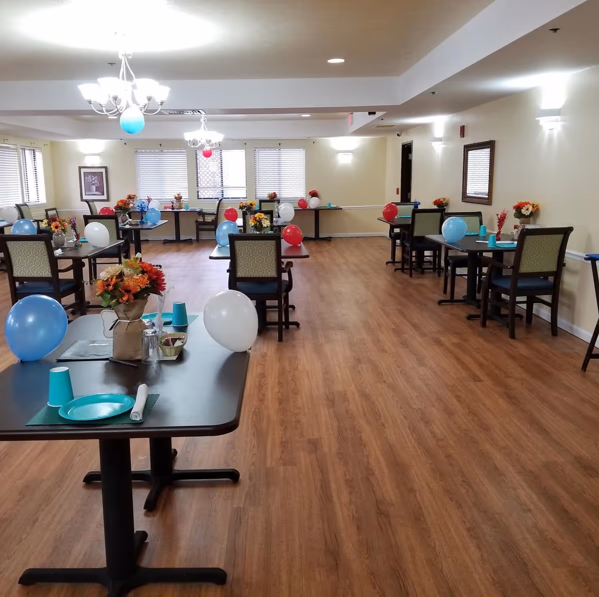 A spacious dining room with several tables and chairs arranged neatly. Each table is decorated with colorful balloons in blue, white, and red, as well as flower centerpieces. The room has wooden flooring, cream-colored walls, and multiple windows with blinds allowing natural light. Ceiling chandeliers and wall sconces provide additional lighting.