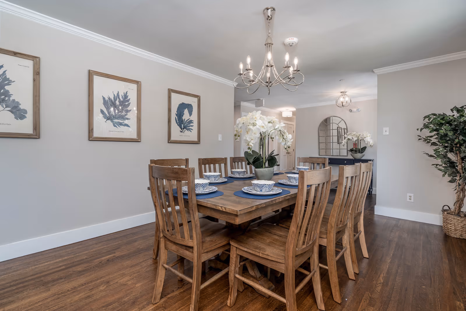 Dining room with a wooden table and eight matching chairs set with blue placemats and a white floral centerpiece under a chandelier.