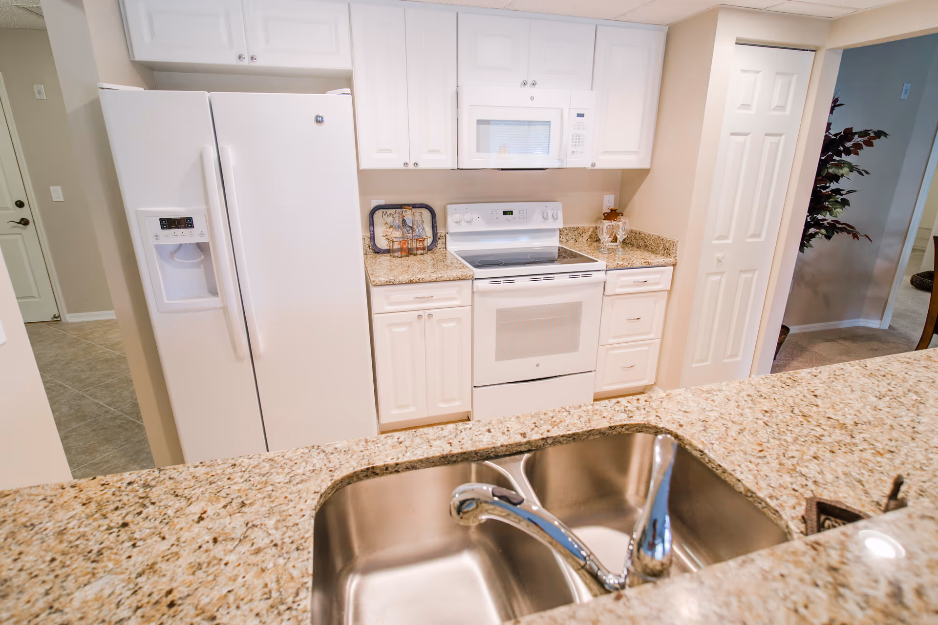 Modern kitchen with white cabinets, a white refrigerator with water dispenser, a white electric stove with oven, a white microwave above the stove, and granite countertops. In the foreground, there is a double stainless steel sink with a chrome faucet. The kitchen opens to an adjacent room with a plant visible.