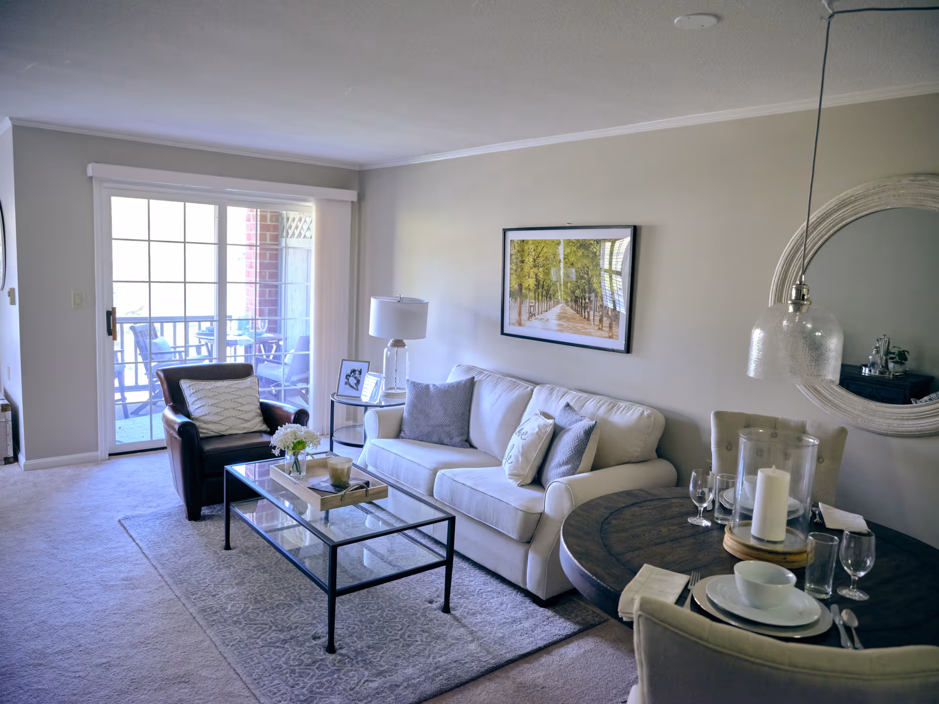 A cozy living room with a white sofa adorned with cushions, a dark armchair with a patterned pillow, a glass coffee table with a flower vase and decorative items, and a round wooden dining table set with plates, glasses, and candles. A large window with sliding glass doors leads to a balcony with outdoor chairs. The walls are light-colored with a framed picture and a round mirror.