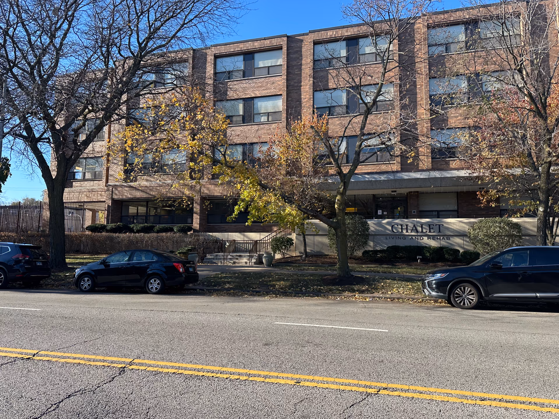 Exterior view of a four-story brick building with several windows, trees with autumn leaves in front, and three parked cars along the street. A sign on the building reads 'CHALET LIVING AND REHAB'.