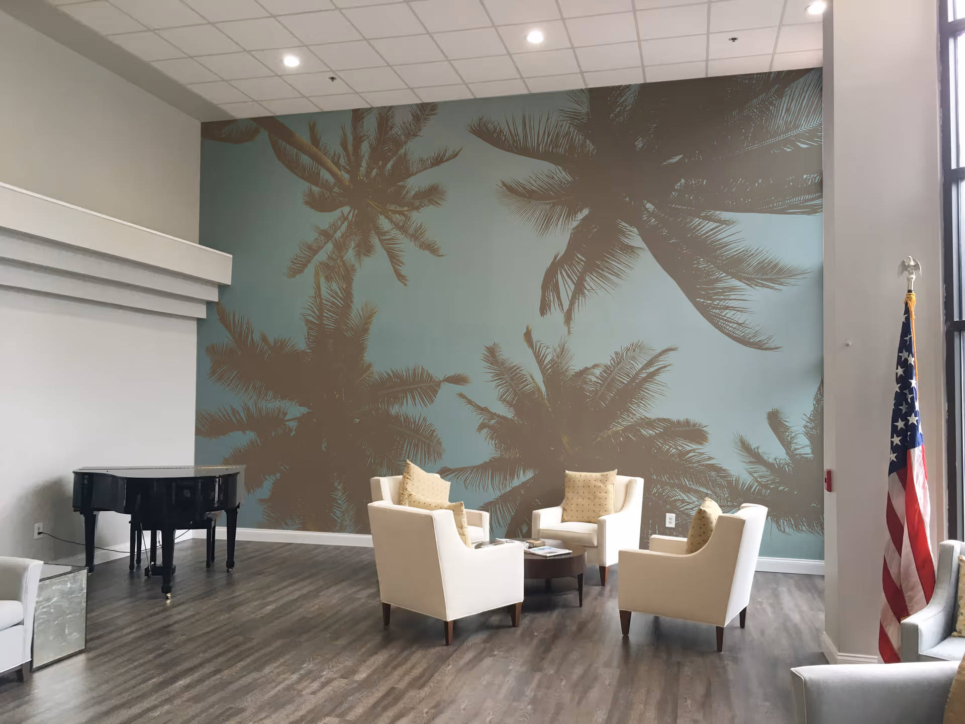 A bright and spacious living room area with four white armchairs arranged around a small round wooden table. The back wall features a large mural of palm tree silhouettes against a blue sky. To the left, there is a black grand piano. On the right side near a large window, an American flag is displayed. The floor is wood, and the ceiling has recessed lighting.