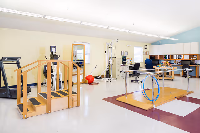 A spacious physical therapy or rehabilitation room with exercise equipment including a treadmill, parallel bars for walking practice, a wooden staircase with handrails, a red exercise ball, and various therapy machines. There are desks and chairs along the back wall with cabinets above them. The walls are painted light yellow and blue.