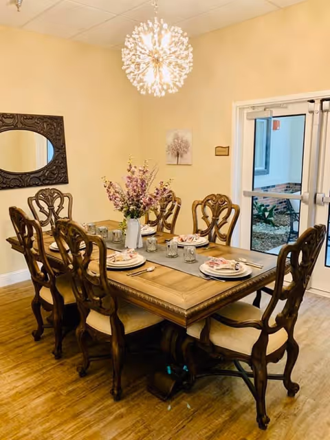 A dining room with a wooden dining table set for six people. The table has a gray runner, plates with napkins, glasses, and a vase with purple flowers in the center. The chairs are wooden with cushioned seats. A decorative mirror hangs on the wall, and a modern chandelier with multiple small lights is above the table. There is a glass door leading outside.