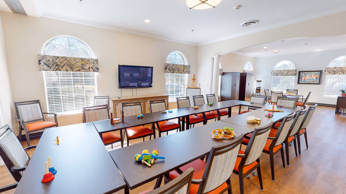 A bright and spacious activity room with large windows featuring arched tops and patterned valances. The room has multiple tables arranged in a U-shape with chairs that have orange cushions and striped backs. Various small musical instruments like tambourines, maracas, and bells are placed on the tables. A flat-screen TV is mounted on the wall above a wooden cabinet. The room has light-colored walls, wooden flooring, and ceiling lights, creating a welcoming atmosphere.