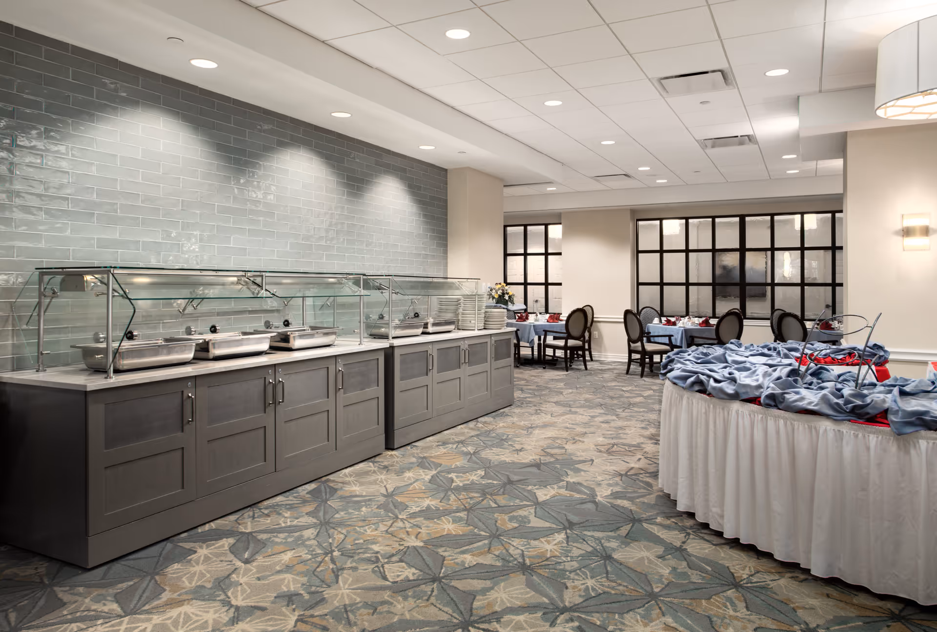 A dining area with a buffet counter featuring covered food trays and plates, tables set with blue tablecloths and chairs in the background, and a round table covered with a white skirt and blue cloth in the foreground. The room has patterned carpet flooring, light-colored walls, and large windows with black frames.