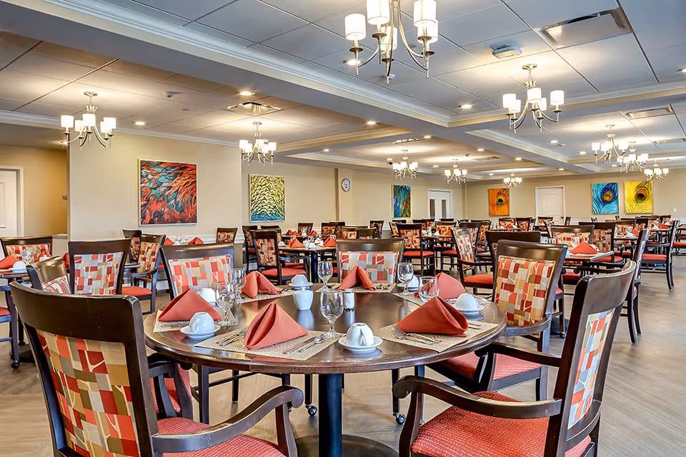 A spacious dining room with multiple round and rectangular tables set with red folded napkins, white cups, glasses, and silverware. The chairs have patterned upholstery in shades of red, beige, and brown. The room is well-lit with multiple chandeliers hanging from the ceiling, and colorful abstract paintings adorn the beige walls.