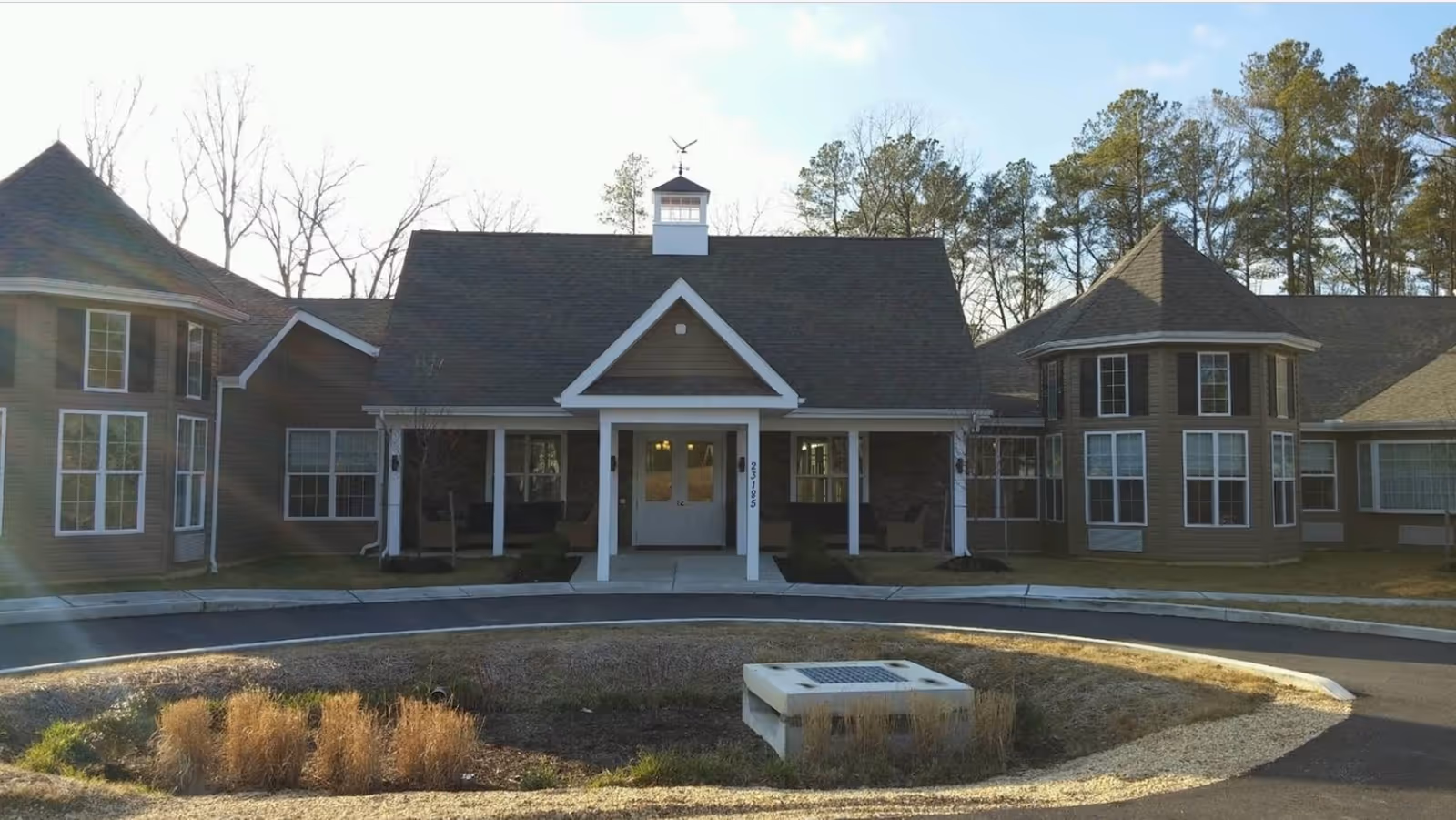Front exterior view of a senior living facility building with a peaked roof, large windows, and a covered entrance. There is a circular driveway with landscaping in front, and trees in the background under a partly cloudy sky.