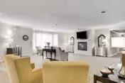 A spacious and well-lit living room with beige carpeting and light-colored walls. The room features two yellow armchairs in the foreground, a fireplace with a mounted TV above it, a mirrored chest of drawers, and a large window with sheer curtains allowing natural light to fill the space. Various decorative items and lamps are placed around the room, creating a cozy and inviting atmosphere.