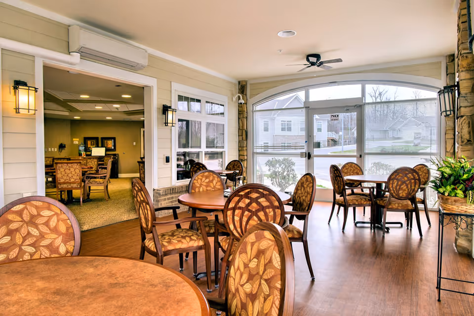 A bright indoor seating area with multiple round wooden tables and chairs featuring leaf-patterned upholstery. Large windows and a glass door allow natural light to fill the space. The room has light-colored walls, wooden flooring, and wall-mounted lantern-style lights. A ceiling fan and air conditioning unit are visible. Through an open doorway, another room with additional seating and framed artwork on the walls can be seen.