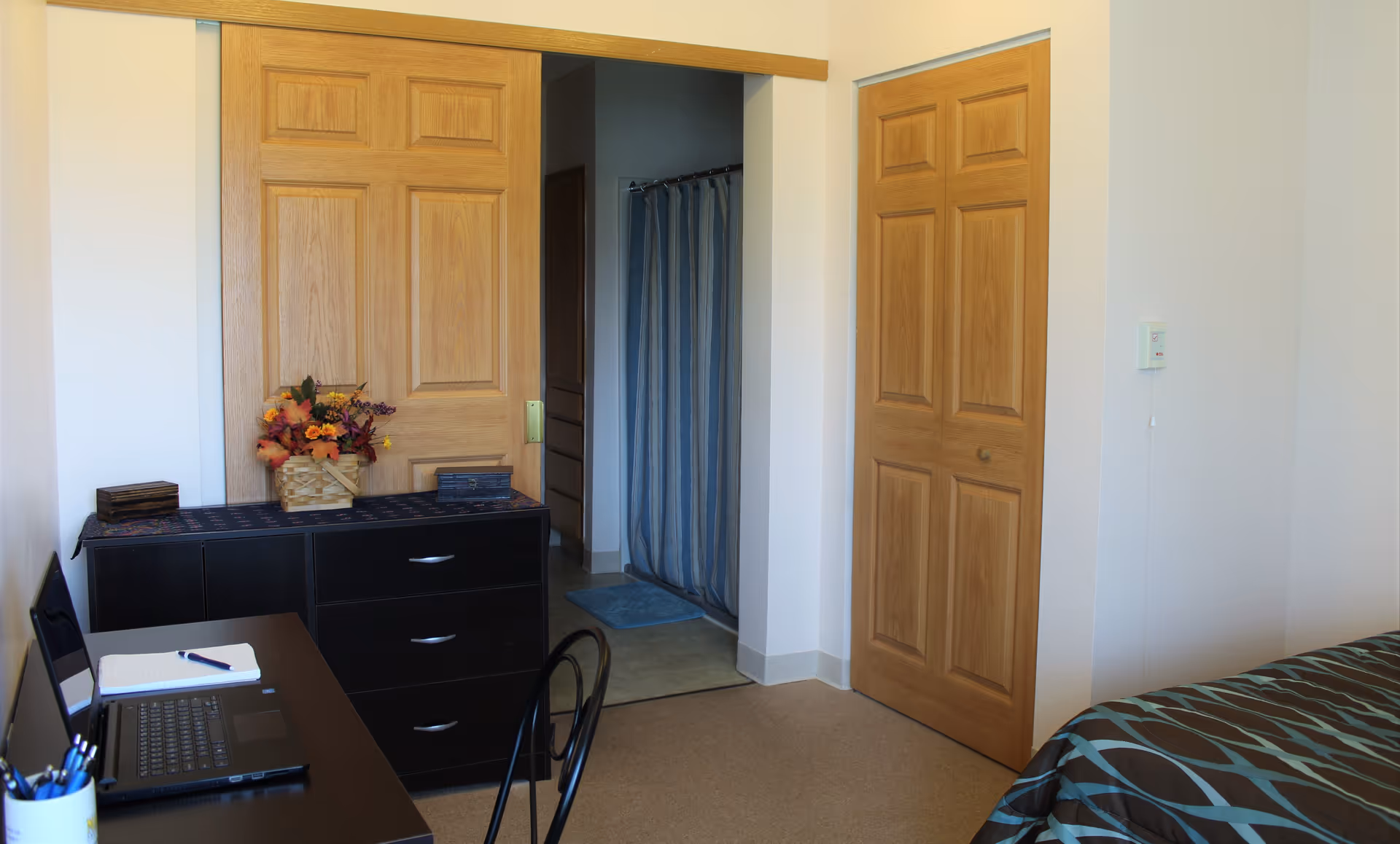 A small bedroom with a bed covered in a brown and blue patterned blanket on the right. There is a black desk with a laptop, notebook, and pen on the left side. A black dresser with a basket of artificial flowers and two small boxes is against the wall. Two wooden doors are visible, one sliding door leading to a bathroom with a blue shower curtain and a blue bath mat, and another closed door on the right wall.