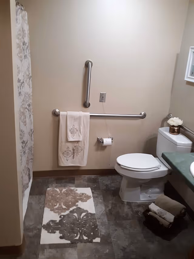 A bathroom with a toilet, a towel rack with two towels, a toilet paper holder with a roll of toilet paper, a decorative rug on the floor, and a shower curtain with a floral pattern. There is also a small basket with rolled towels on the floor next to the toilet and a small flower arrangement on the toilet tank.
