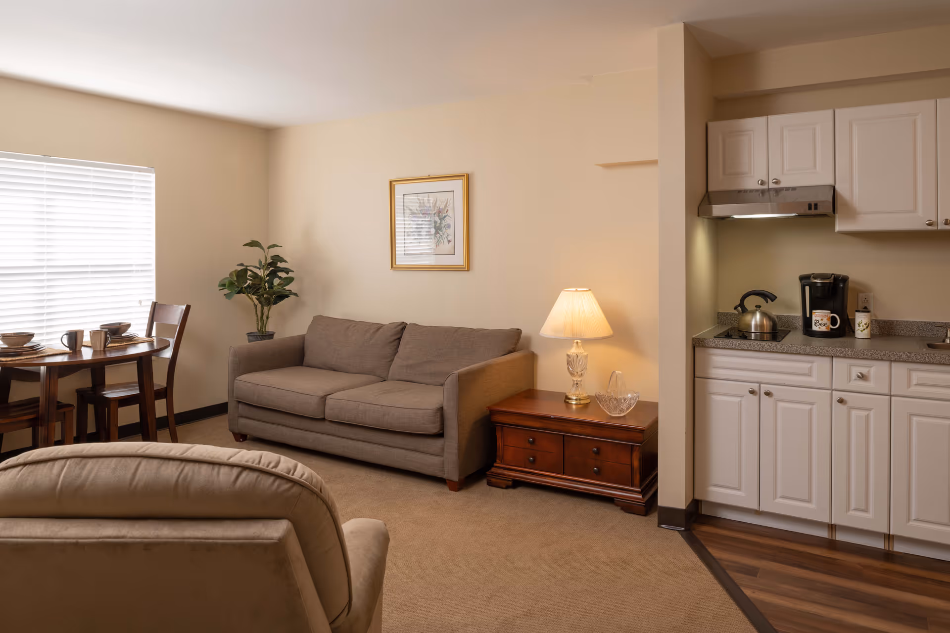 A cozy living area in a senior living facility featuring a beige sofa, a wooden side table with a lamp and a glass bowl, a small dining table with two chairs set with dishes and cups, a potted plant, and a kitchenette with white cabinets, a kettle, and a coffee maker.