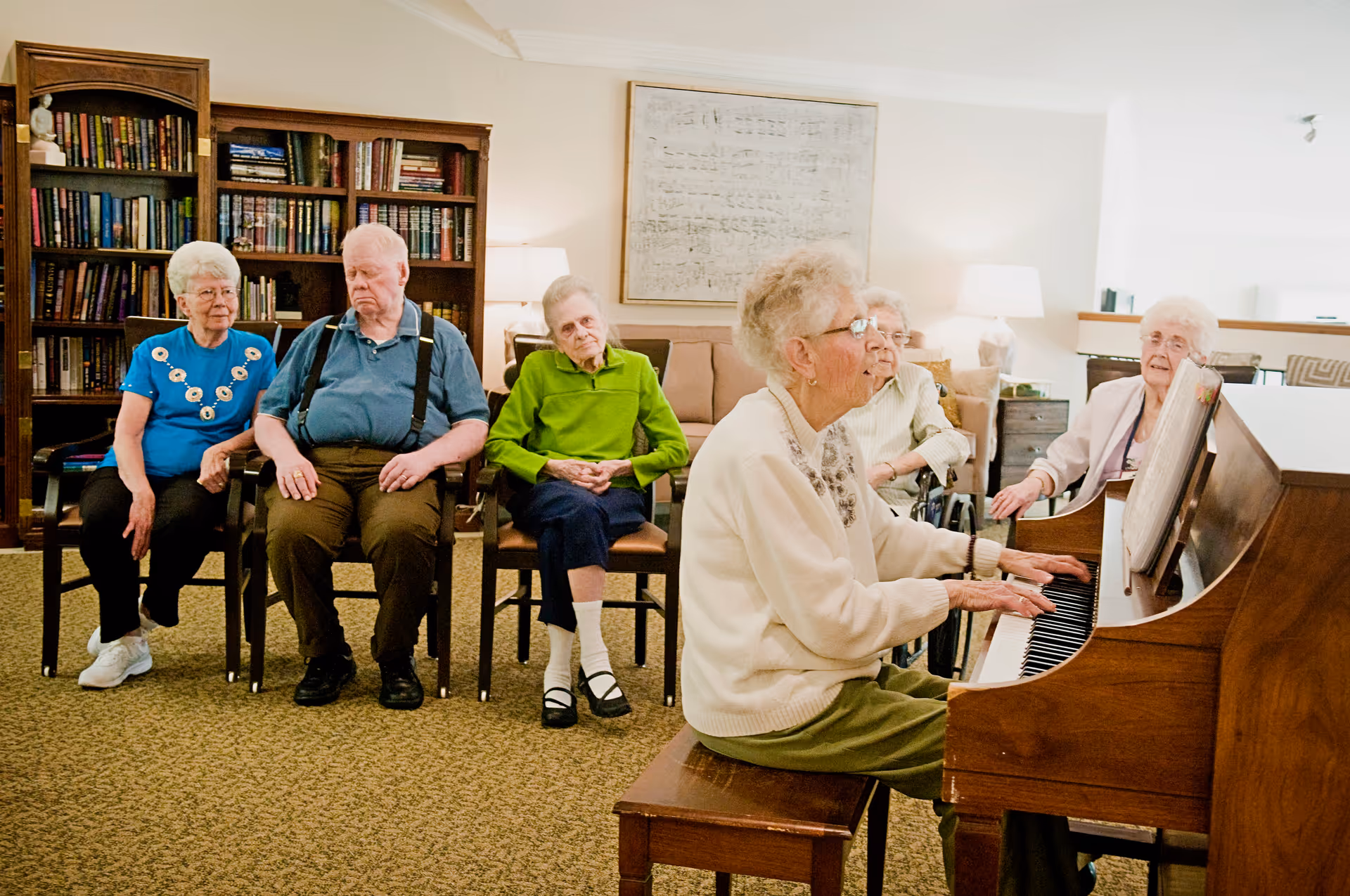 A group of elderly people sitting in chairs in a living room area, with one elderly woman playing a piano. The room has a bookshelf filled with books, a couch, lamps, and framed artwork on the wall.