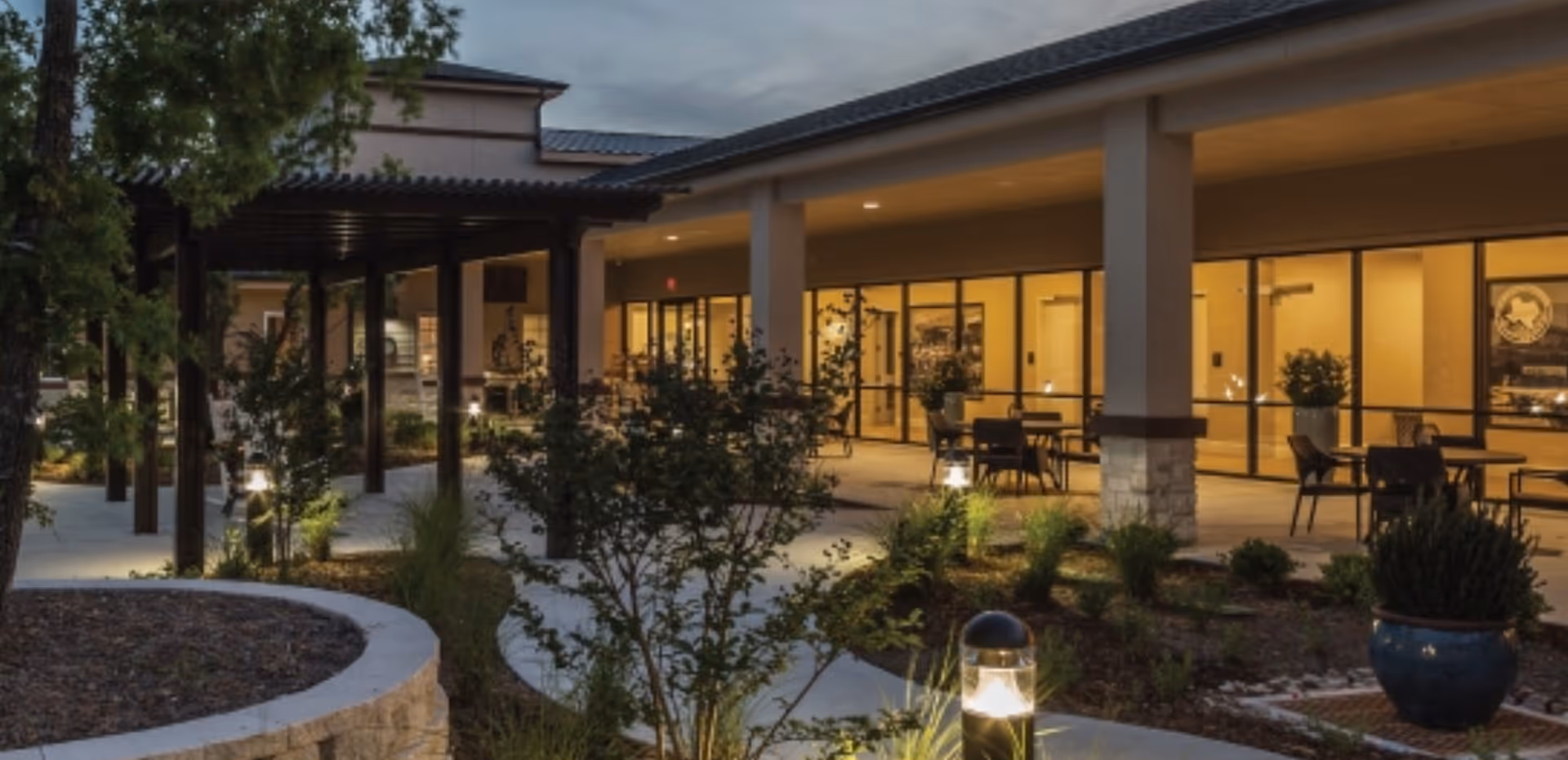 Evening view of an outdoor patio area at a senior living facility with tables and chairs under a covered walkway, surrounded by landscaped plants and illuminated by pathway lights.