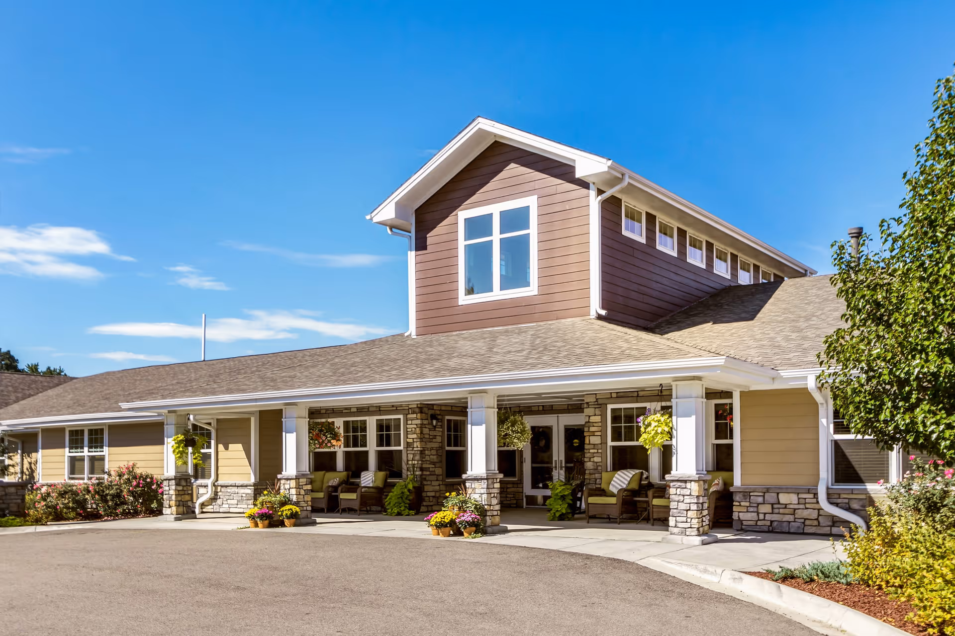 Exterior view of a single-story senior living facility building with a covered entrance supported by stone pillars, outdoor seating, and hanging plants under a clear blue sky.
