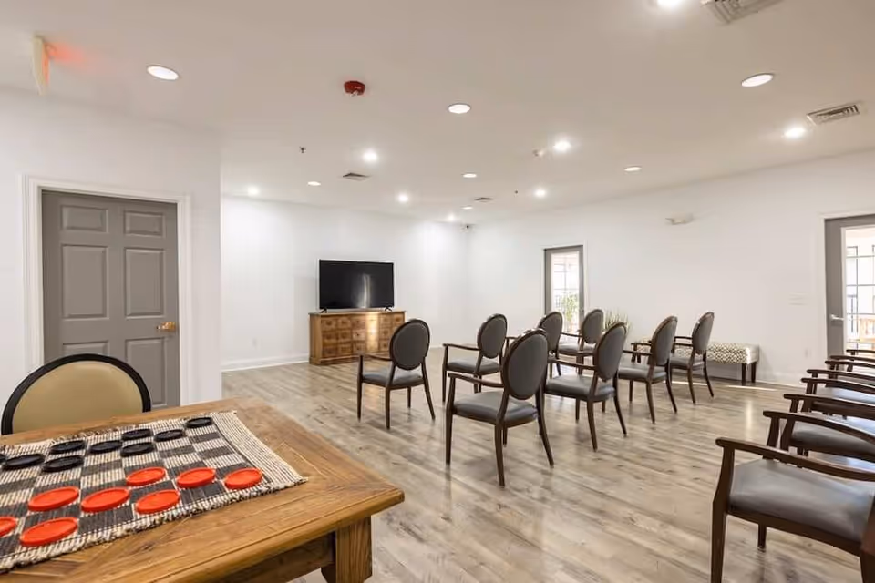 A bright, spacious room with wooden flooring and white walls, furnished with multiple chairs arranged in rows facing a flat-screen TV on a wooden cabinet. In the foreground, there is a table with a checkers game set up. The room has two doors and a window, with recessed ceiling lights providing illumination.