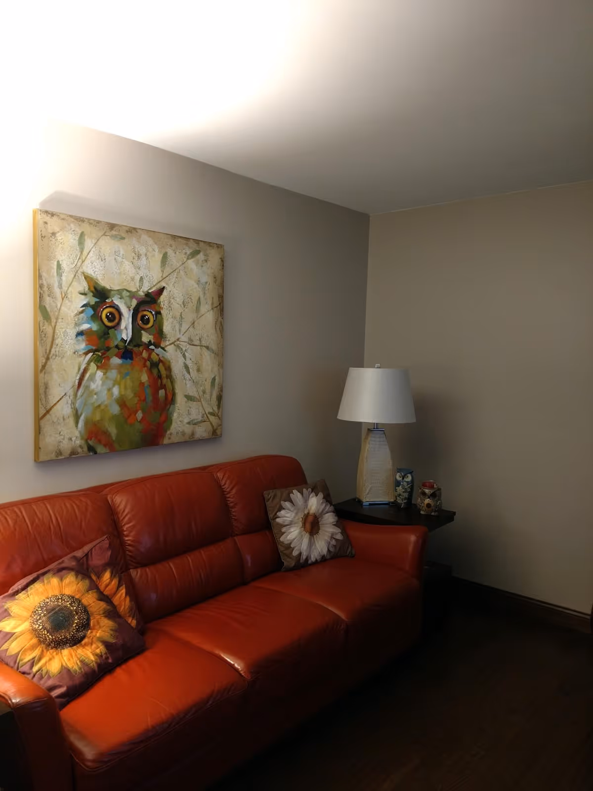 A cozy living room with a red leather couch adorned with two decorative pillows featuring sunflower and daisy designs. Above the couch hangs a colorful painting of an owl. To the right of the couch is a small side table with a lamp and decorative items. The walls are painted beige and the floor is dark wood.