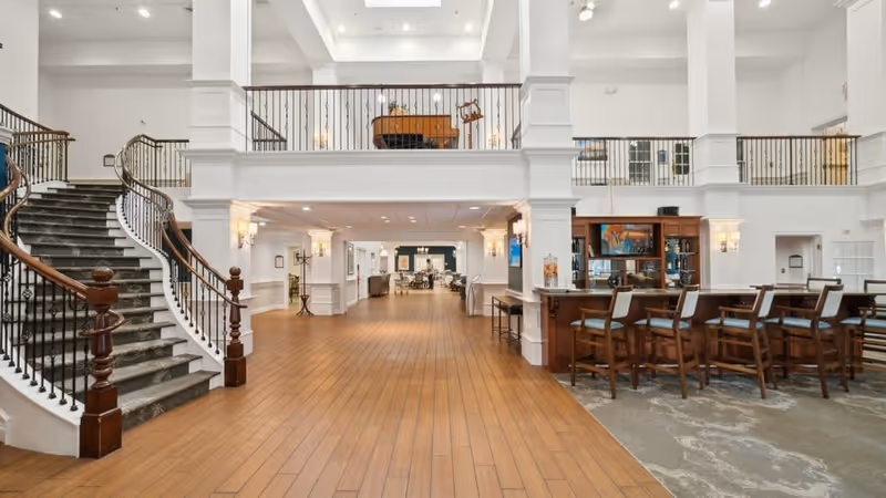 Spacious two-story senior living lobby with a grand curved staircase, wood floors, and a bar seating area.