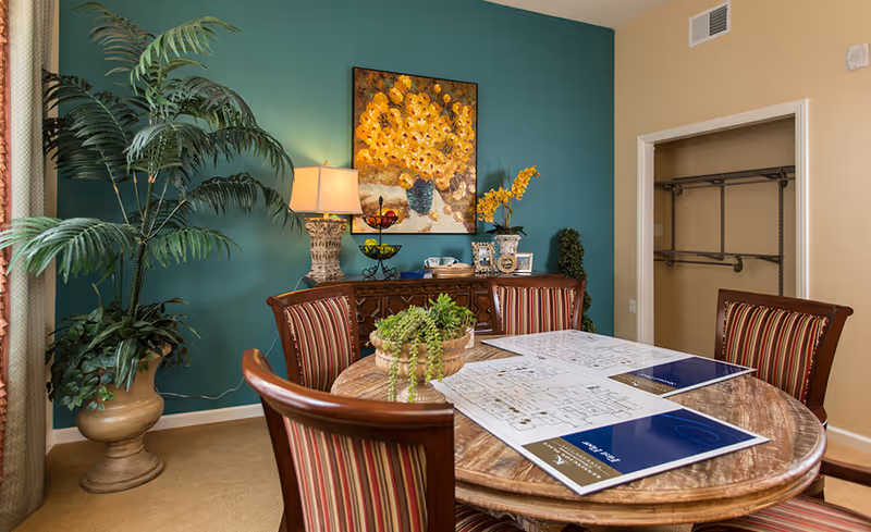 Round wooden dining table with striped chairs and floor plans on it in a decorated room with a teal accent wall, artwork, lamp, and potted plants.