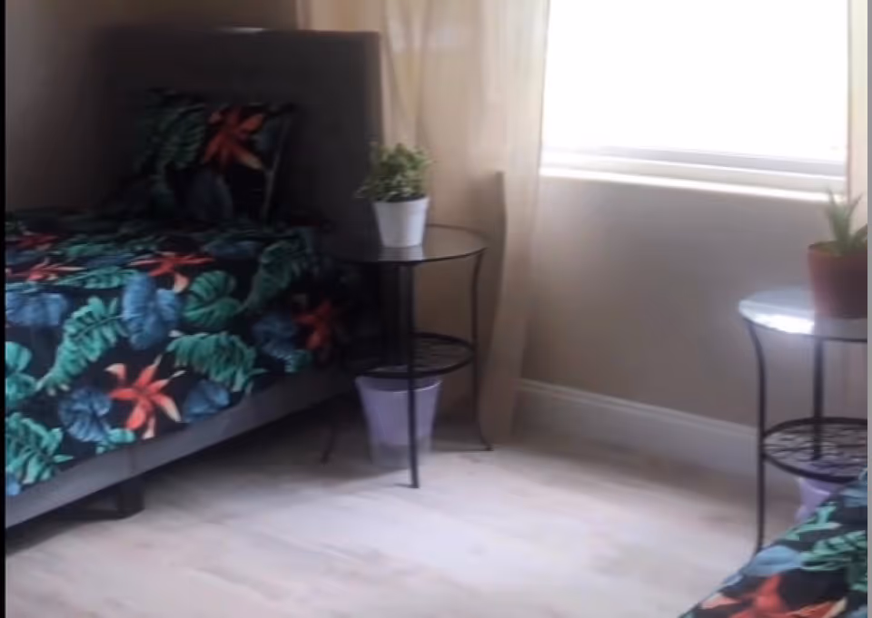 A bedroom with two beds featuring dark floral-patterned bedding. Each bed has a small round black metal side table next to it, with a potted plant on top. There is a window with light-colored curtains allowing natural light into the room.