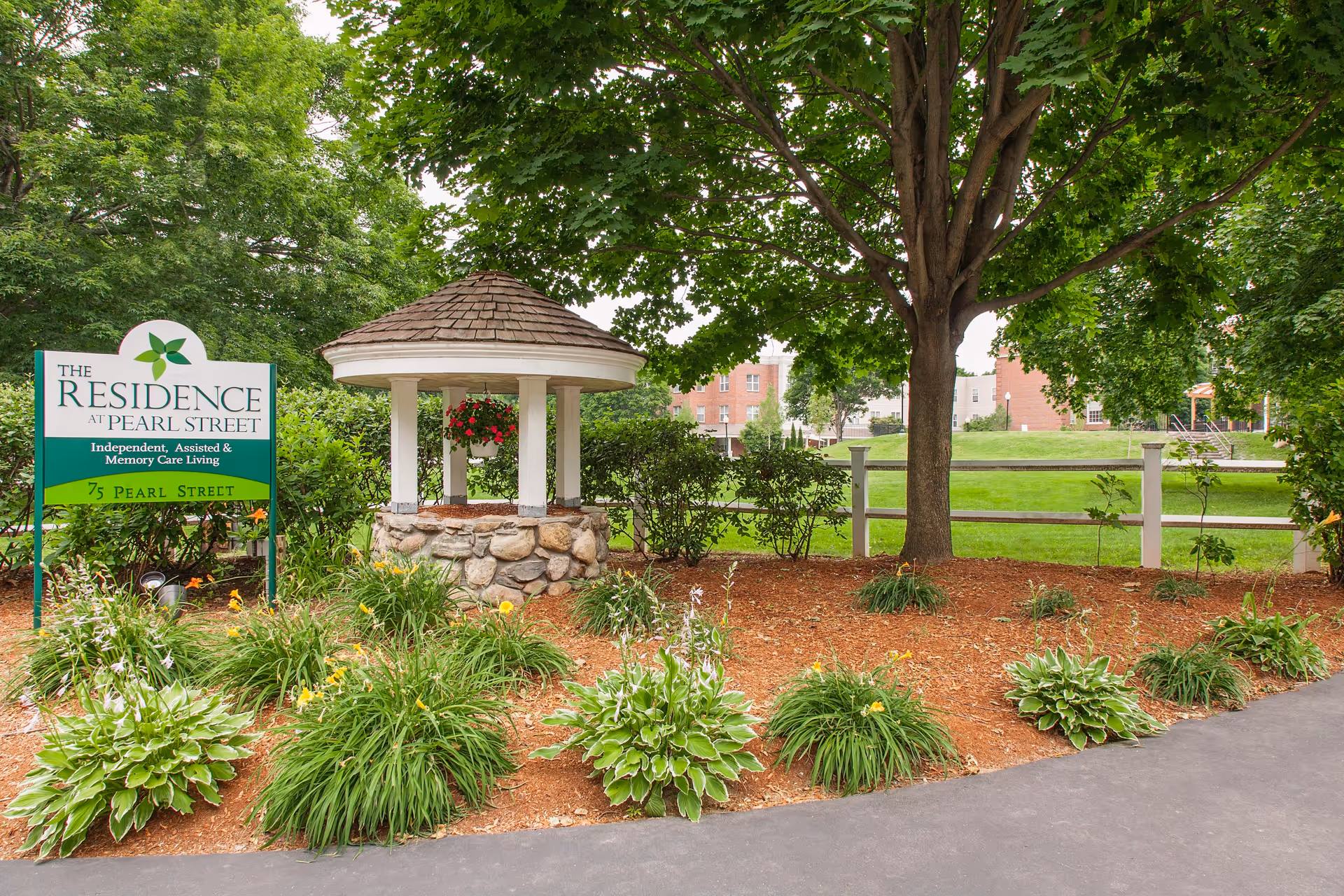 Outdoor garden area at The Residence at Pearl Street featuring a stone well with a shingled roof, surrounded by green plants and flowers, with a large tree providing shade and a white fence in the background.