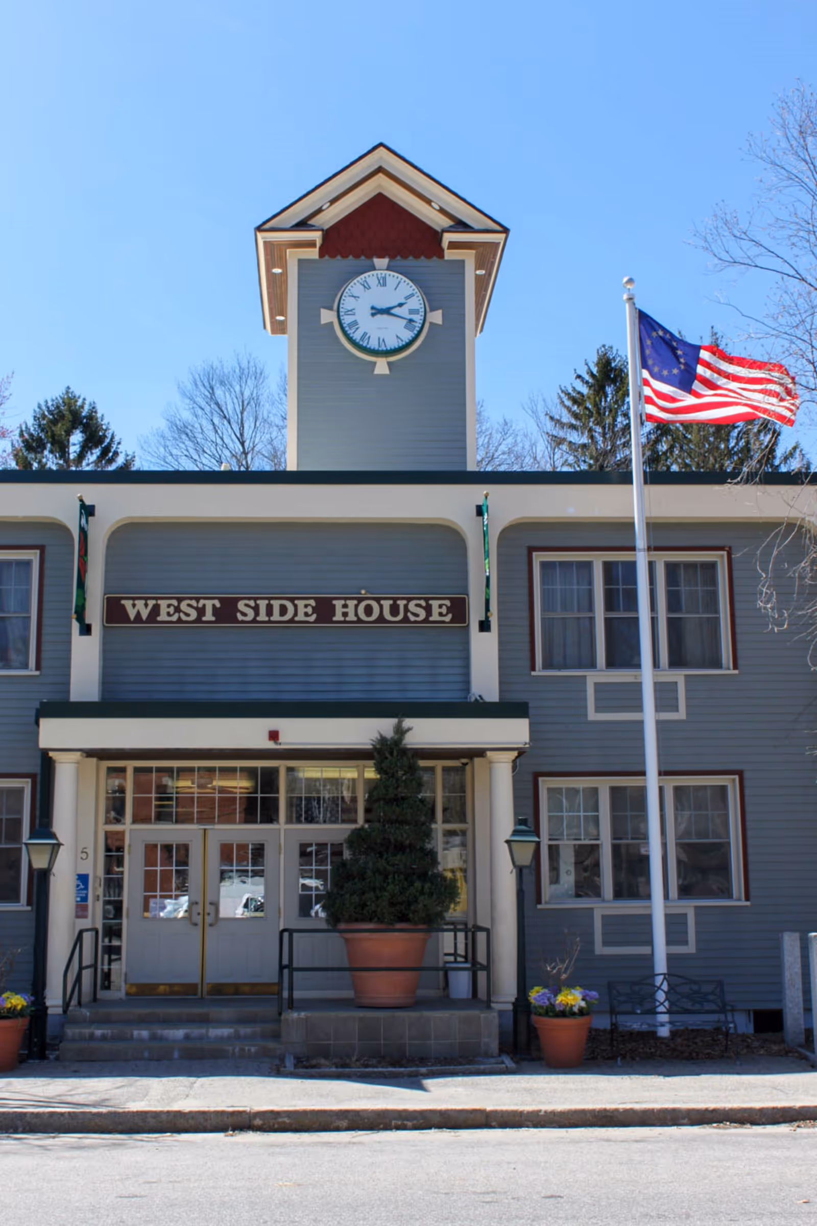 Front exterior view of the West Side House building with a clock tower above the entrance, an American flag on a flagpole to the right, potted plants near the entrance, and clear blue sky in the background.