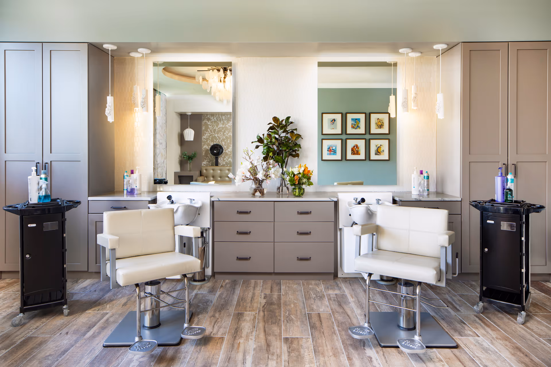 Interior view of a senior living facility's hair salon area with two white salon chairs in front of two washbasins and large mirrors. The space features wood-look flooring, beige cabinetry, two black rolling carts with hair products, and decorative plants and flowers on the counter between the washbasins.