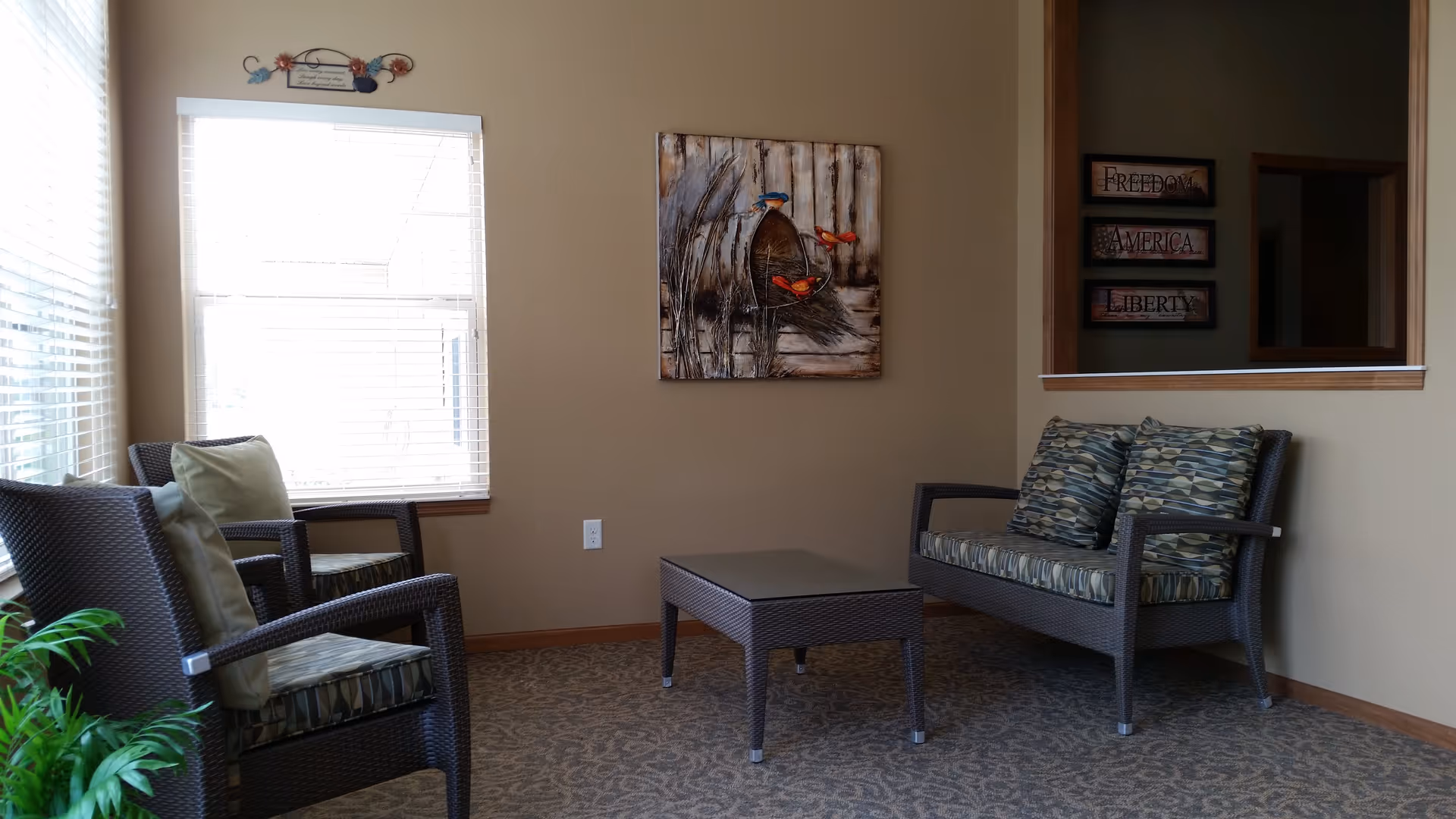 A cozy sitting area with two wicker armchairs and a wicker loveseat, all with patterned cushions. A small rectangular wicker coffee table is centered between the seating. The walls are beige with a decorative painting of birds and a basket hanging on one wall. There is a window with blinds letting in natural light and a small plant in the corner.