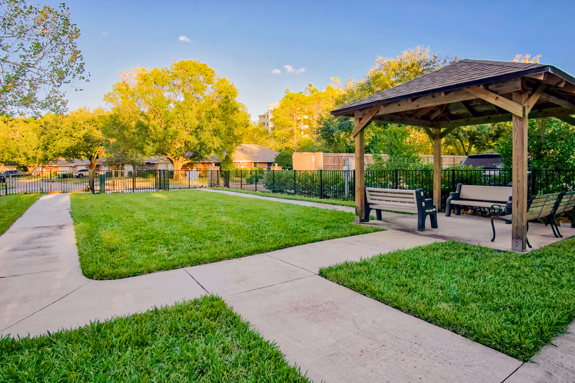 A grassy courtyard with paved walkways, a wooden gazebo and benches surrounded by trees and a fence.
