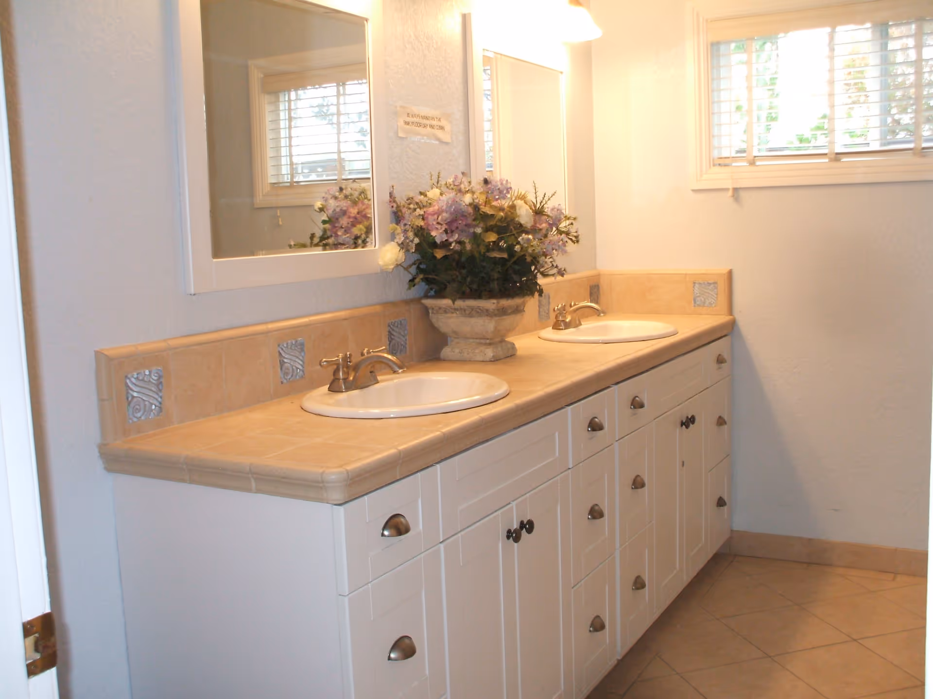 A bathroom vanity with two sinks, beige tiled countertop, and white cabinetry. Above the sinks are two mirrors and a decorative floral arrangement placed between them. A window with white blinds is visible on the right wall.