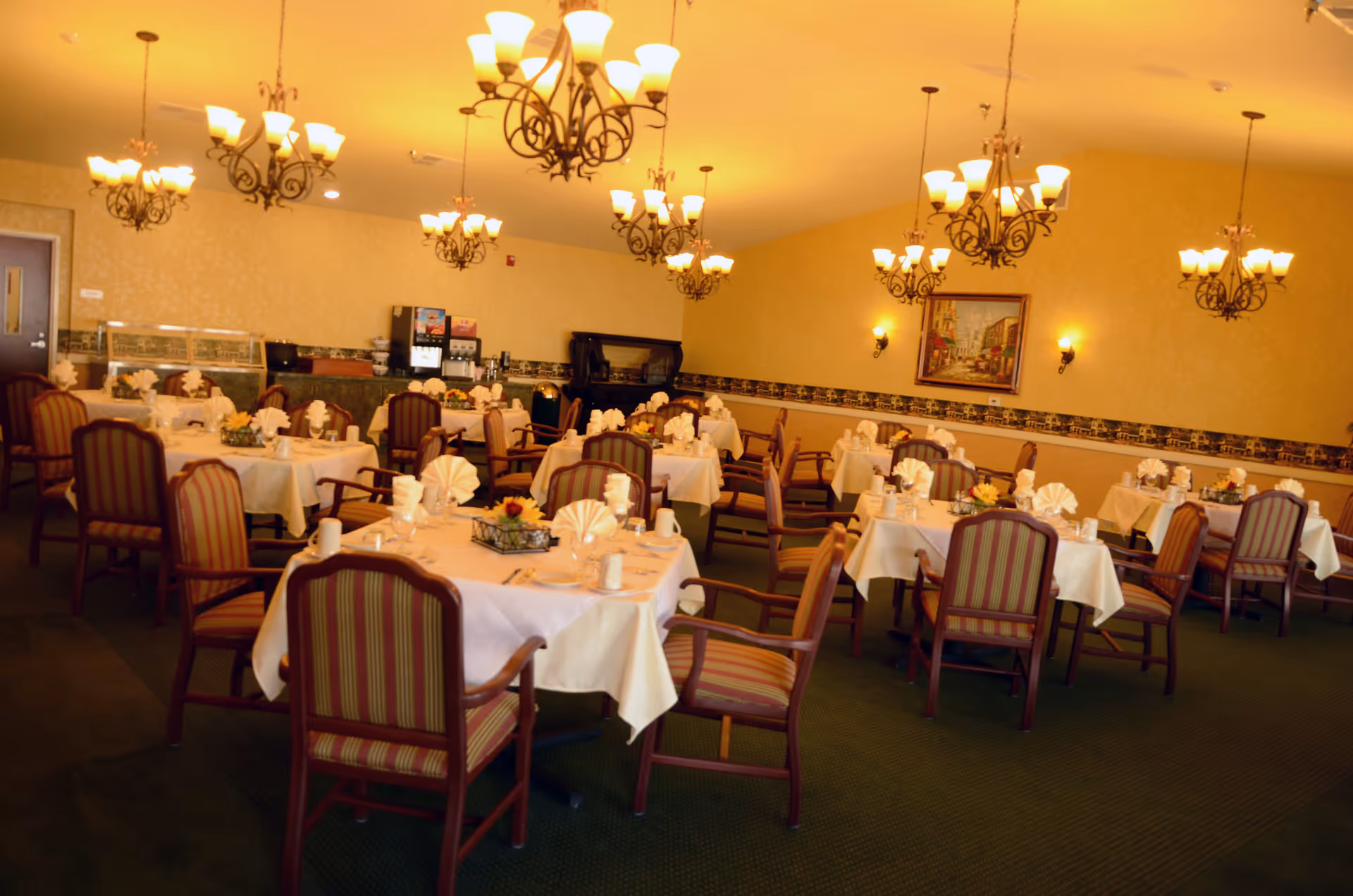 A warmly lit dining room with multiple tables covered in white tablecloths, each set with folded napkins, cups, and utensils. The room features striped upholstered chairs, ornate chandeliers hanging from the ceiling, a painting on the wall, and a beverage station in the back.