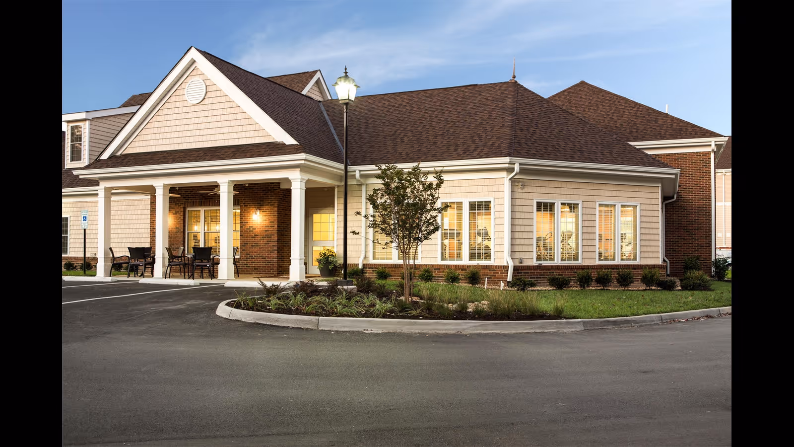 Exterior view of a single-story building with beige siding and brick accents, featuring a covered porch with chairs and tables. The building is surrounded by a small landscaped area with grass, shrubs, and a tree, and a street lamp is visible near the entrance. The sky is clear and blue.