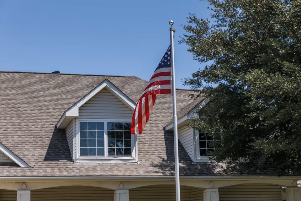 Exterior view of a building with a gray shingled roof and white siding, featuring two dormer windows. In front of the building, an American flag is flying on a flagpole. There is a large tree to the right side of the image under a clear blue sky.