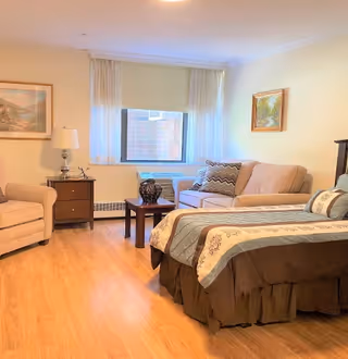 Bright furnished studio-style room with a bed, sofa, armchair, side tables, and a window.