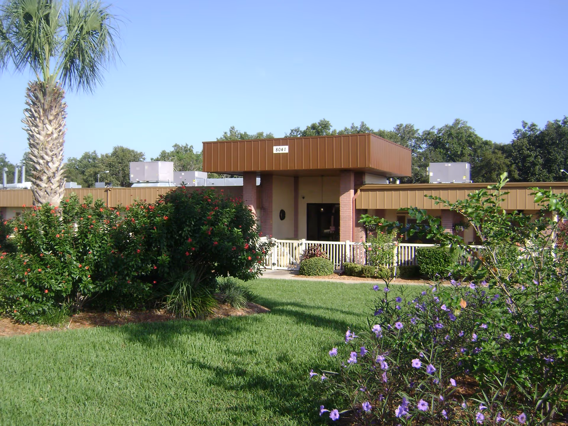 Exterior view of Bear Creek Nursing Center showing a single-story building with a brown roof and entrance canopy. The foreground features a well-maintained lawn, various bushes with red and purple flowers, and a palm tree on the left side. The sky is clear and blue.