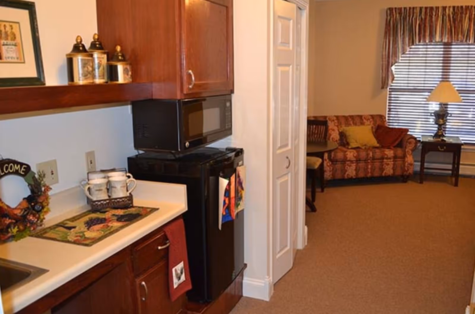 Small kitchenette with a microwave and mini-fridge opening into a living area with a patterned sofa and lamp.