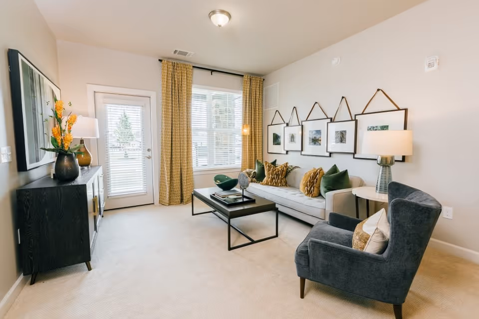Bright living room with a gray sofa, navy armchair, coffee table, gallery of framed photos, and a windowed door with yellow curtains.