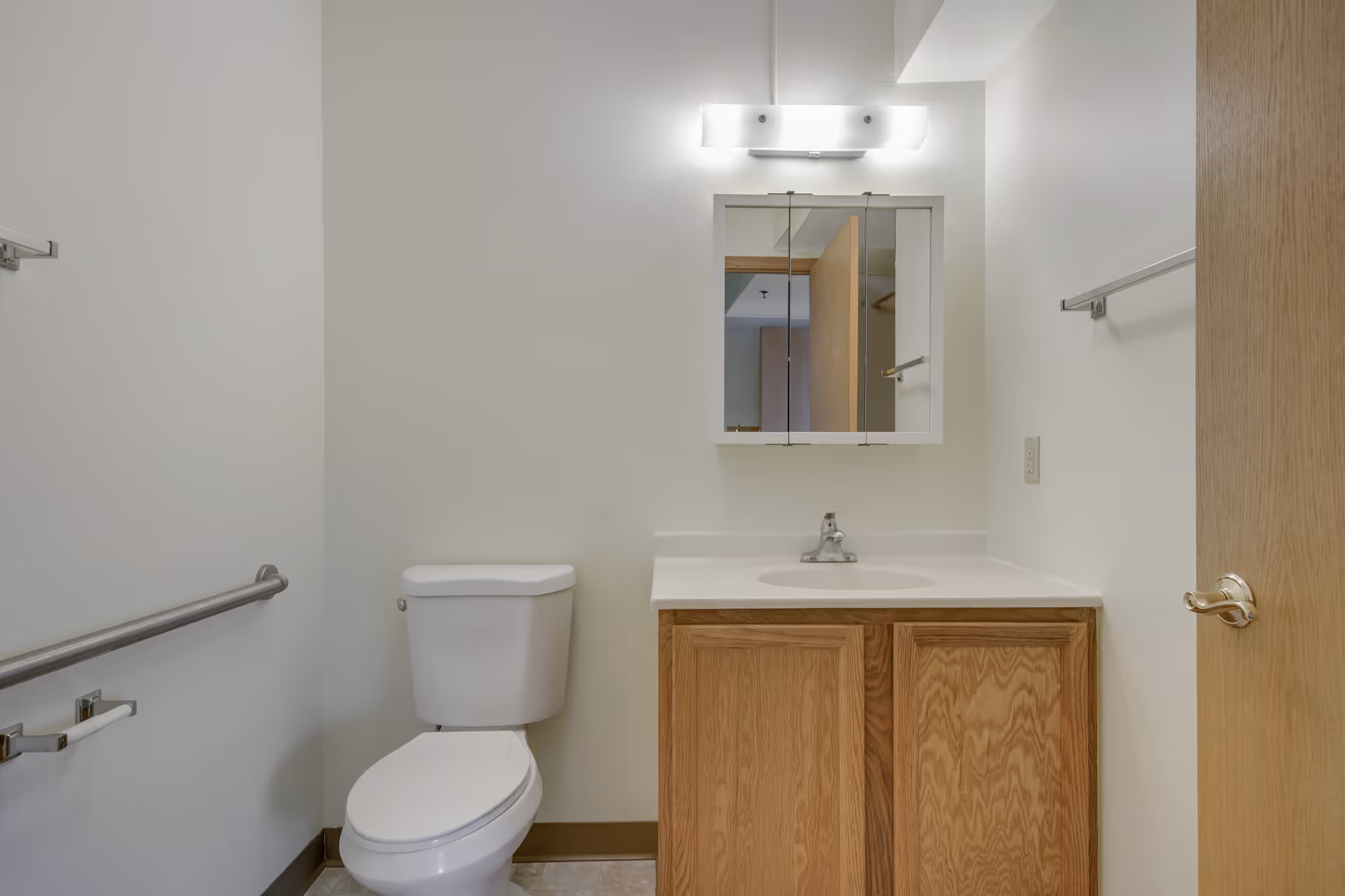A clean bathroom with a white toilet, a wooden vanity with a white countertop and sink, a mirrored medicine cabinet above the sink, a light fixture above the cabinet, and grab bars on the walls.