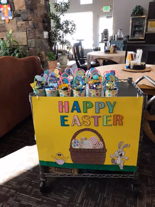 A decorated cart with a bright yellow sign that reads 'HAPPY EASTER' in colorful letters. The sign features a basket filled with decorated Easter eggs, a cartoon chick, and a bunny. On top of the cart are small colorful Easter baskets filled with treats. The cart is in a cozy room with tables, chairs, plants, and a stone fireplace in the background.