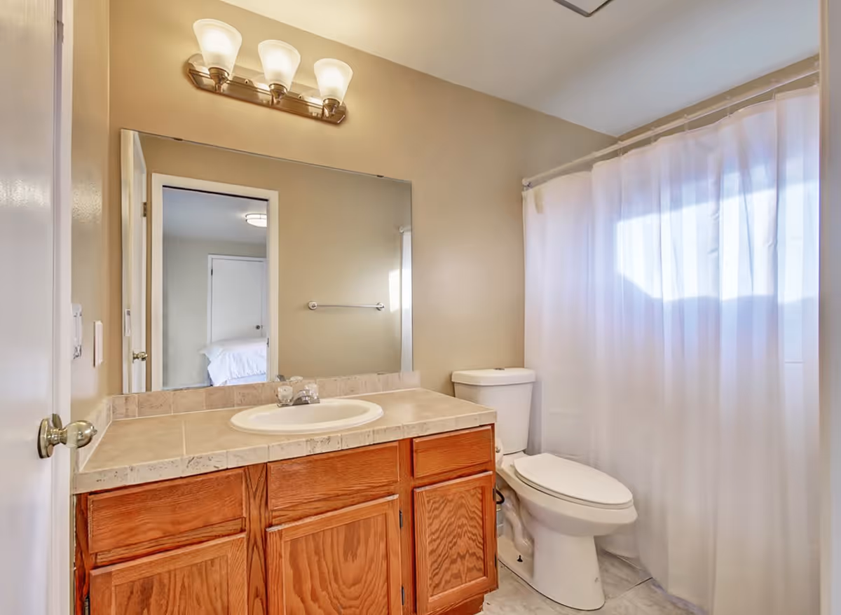 A bathroom with a wooden vanity cabinet topped with a beige tiled countertop and a white sink. Above the sink is a large mirror reflecting a bedroom doorway. To the right of the vanity is a white toilet next to a bathtub with a white shower curtain. The walls are painted beige and there is a three-light fixture above the mirror.