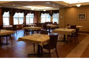 A dining room with multiple tables covered with white tablecloths and set with glasses and silverware. The room has large windows with dark curtains, wooden chairs, and warm lighting from ceiling fixtures.
